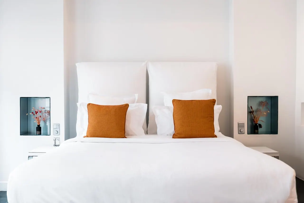A neatly made double bed with white linens and orange accent cushions is set against a light wall.