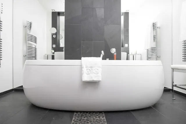 A bright modern bathroom features a freestanding bathtub, dark tiled wall and twin vanity areas.