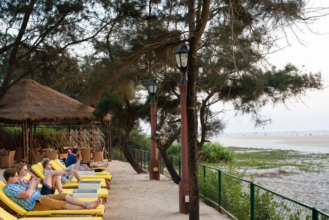A beachfront area has yellow loungers, a thatched dining shelter, decorative lamps and views across the sandy tidal shore.