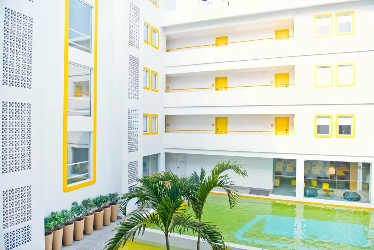 A bright courtyard pool is surrounded by white and yellow hotel buildings and potted palms.