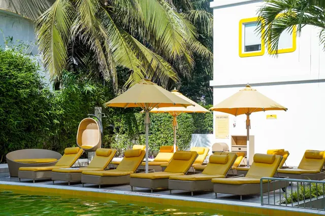 A pool is lined with yellow-cushioned loungers and umbrellas beside tropical greenery.