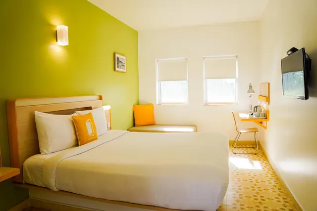 A bright hotel bedroom has a double bed, desk, seating area, television and two windows.