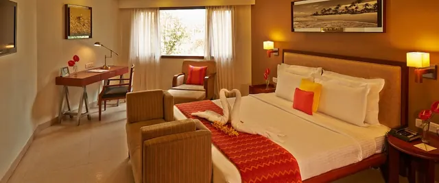 A warmly lit bedroom contains a large bed, seating, a desk, and framed wall art.