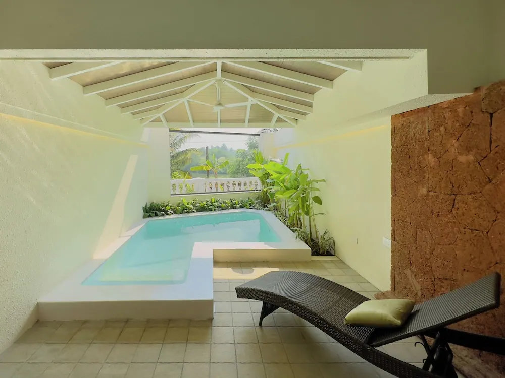 A covered private pool area has a small swimming pool, a wicker lounger, tropical plants and greenery beyond the balustrade.
