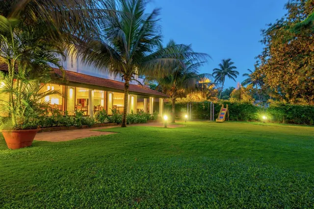 A landscaped tropical garden with palm trees, lawn, lighting and a covered building veranda is shown at dusk.