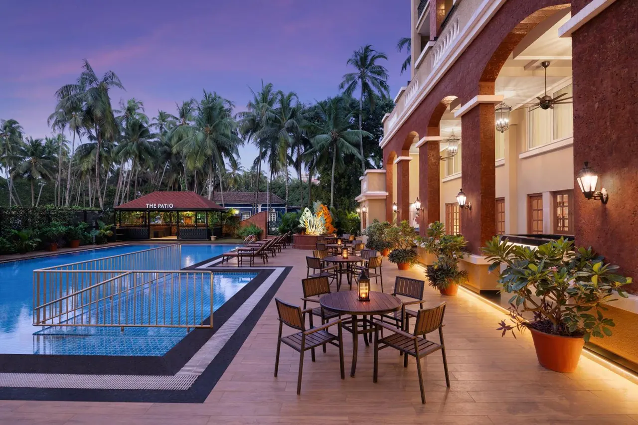A large pool and outdoor dining terrace sit beside an arched tropical hotel building at dusk.