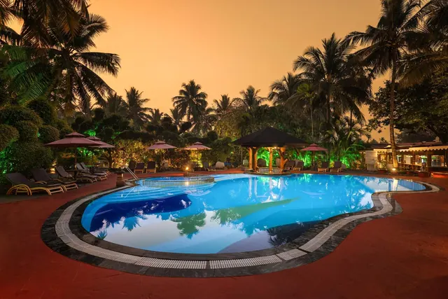 A large illuminated swimming pool is surrounded by sun loungers, umbrellas, tropical palms and a pool bar at sunset.