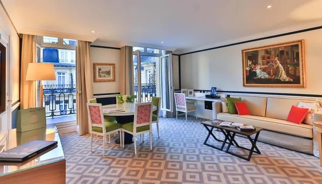 A spacious living area has a sofa, dining table, desk, patterned carpet and tall windows overlooking the street.