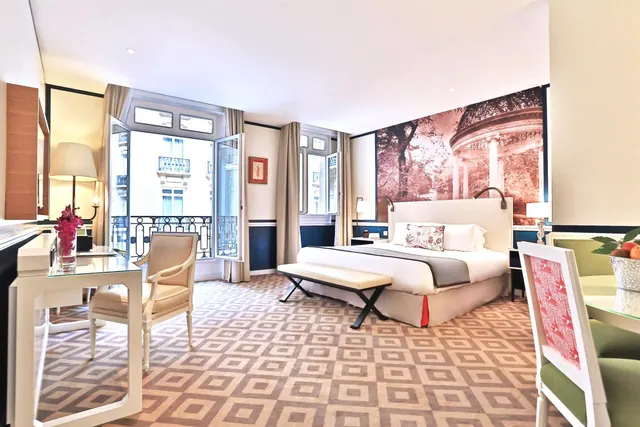 A bright hotel bedroom has a double bed, desk, seating, patterned carpet and open windows overlooking Parisian buildings.