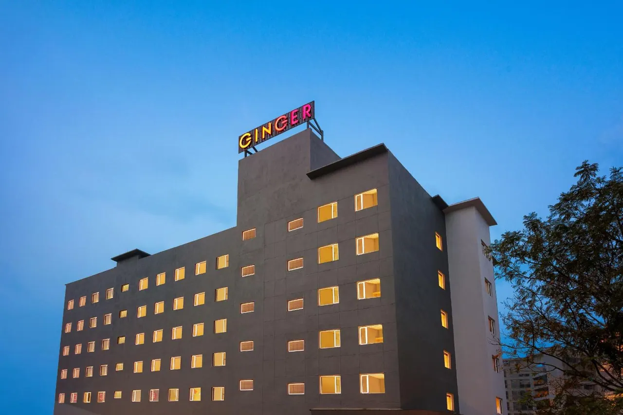 A modern hotel building with illuminated windows and a rooftop GINGER sign is shown against the evening sky.
