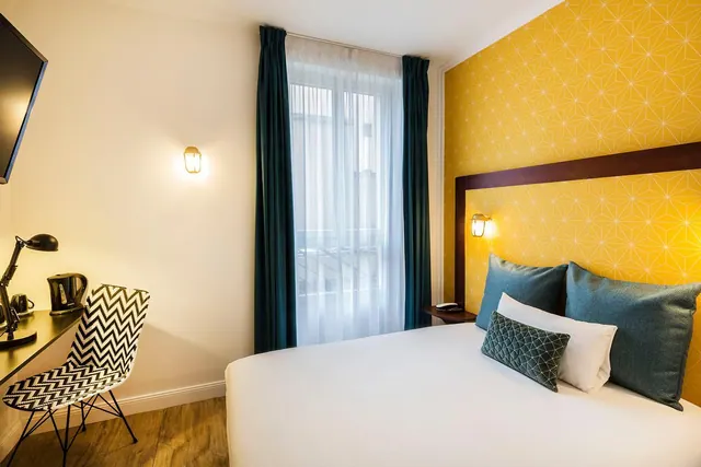 A bright hotel bedroom contains a large bed, desk, kettle, television, patterned accent wall, and tall curtained window.