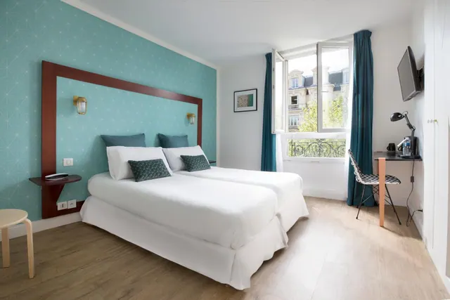 A bright hotel bedroom has a double bed, turquoise patterned wall, work desk, television, and a window overlooking Parisian buildings.