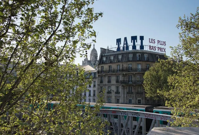 A Parisian streetscape shows a heritage building, a metro bridge, and the Sacré-Cœur Basilica behind the trees.