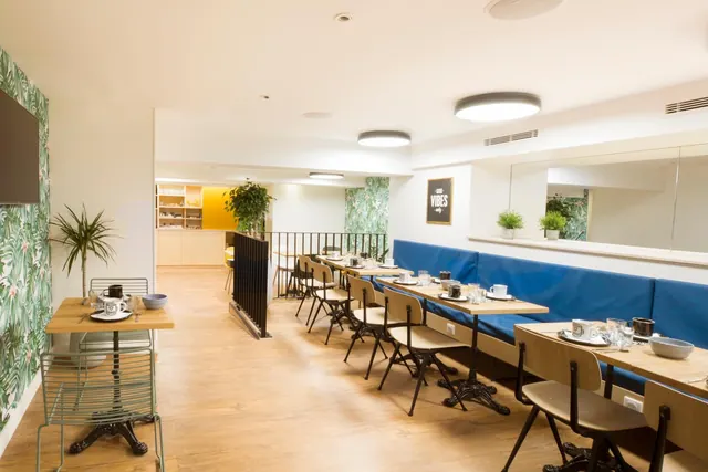A bright dining room has long tables, blue banquette seating, and potted plants.