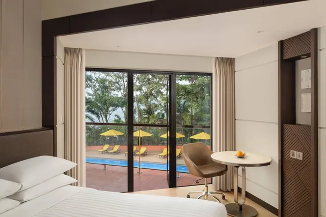 A bright bedroom with a double bed, desk, seating, and views of the pool and landscaped grounds through the glass doors.