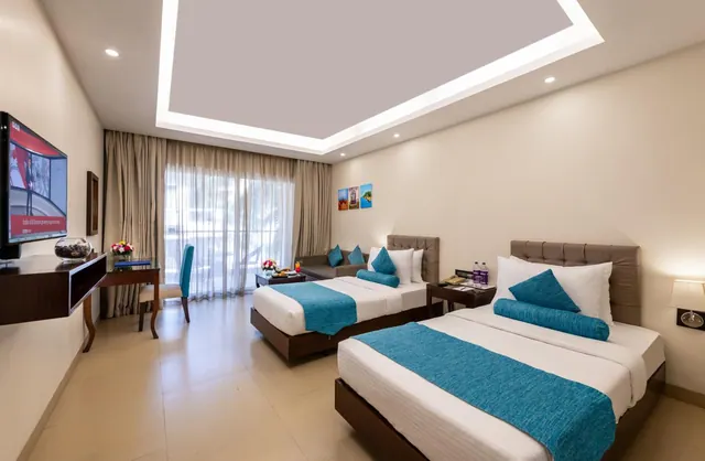 A bright bedroom contains two neatly made beds, a desk, seating, television and full-height curtained windows.