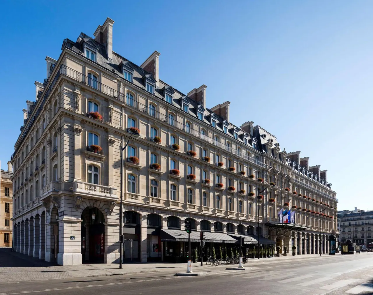 A grand historic hotel building stands along a broad Parisian street under a clear blue sky.
