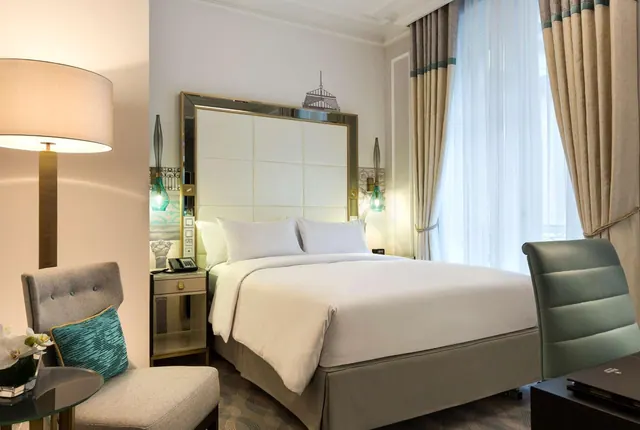 A well-appointed bedroom has a large bed, upholstered seating, a desk, and tall curtained windows.