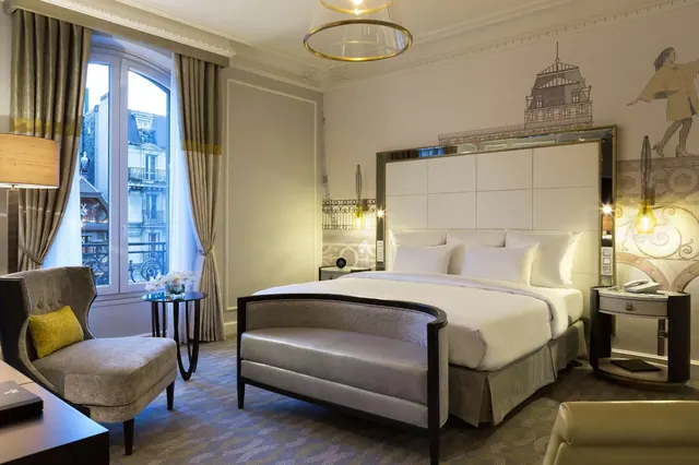 A spacious hotel bedroom has a large bed, a curved sofa, an armchair, and tall windows overlooking the street.