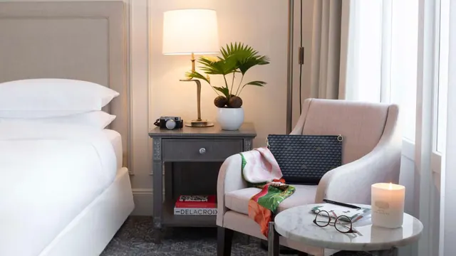 A well-appointed bedroom contains a large bed, an armchair, a bedside table, and a round table beside tall windows.