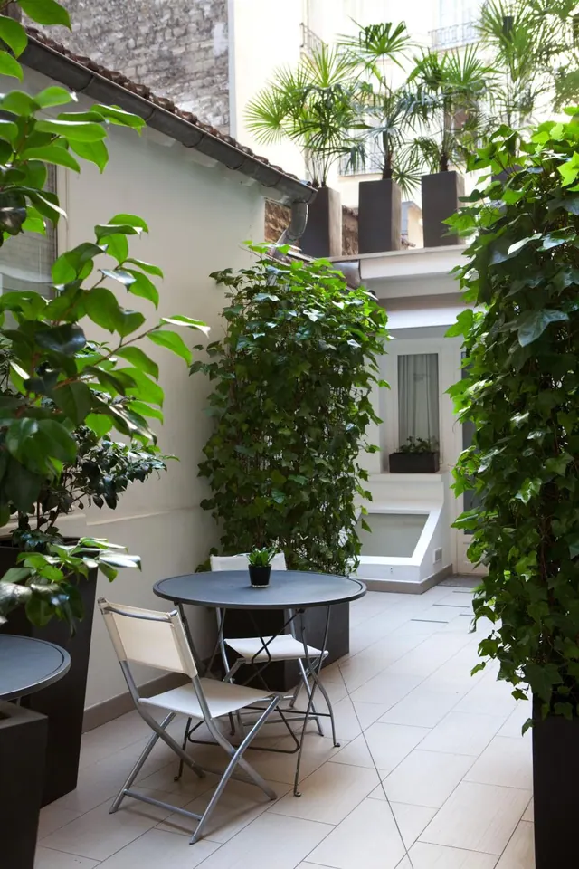 A leafy courtyard contains potted plants, a small café table with folding chairs, and surrounding hotel buildings.