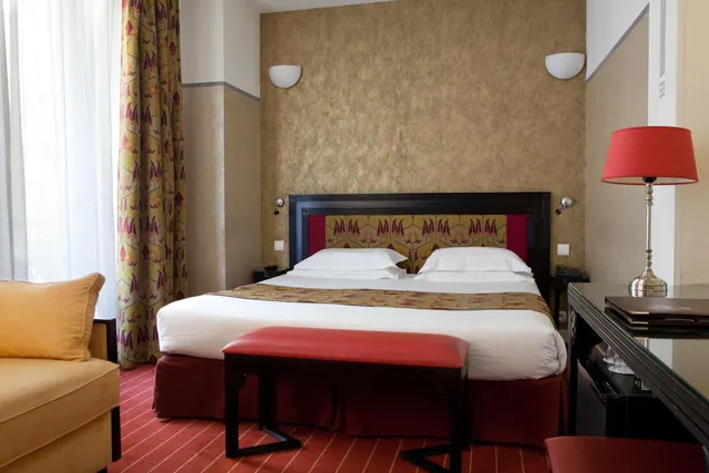 A furnished hotel bedroom with a double bed, seating, patterned curtains, and a large window.