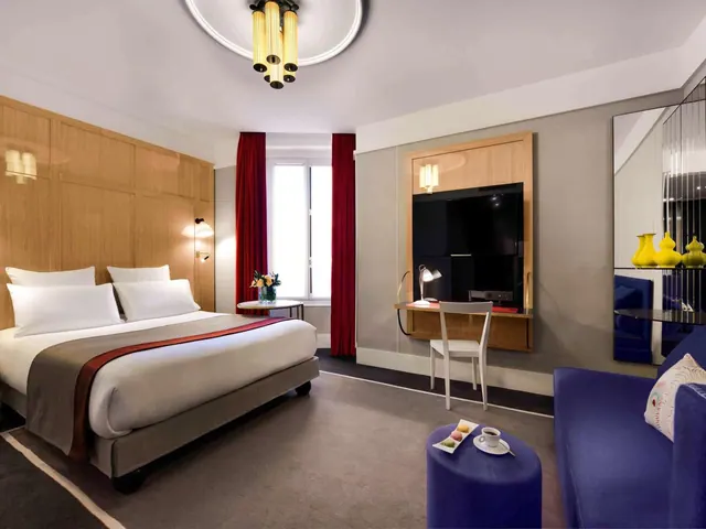 A stylish hotel bedroom contains a double bed, desk, television, wardrobe and blue seating area.