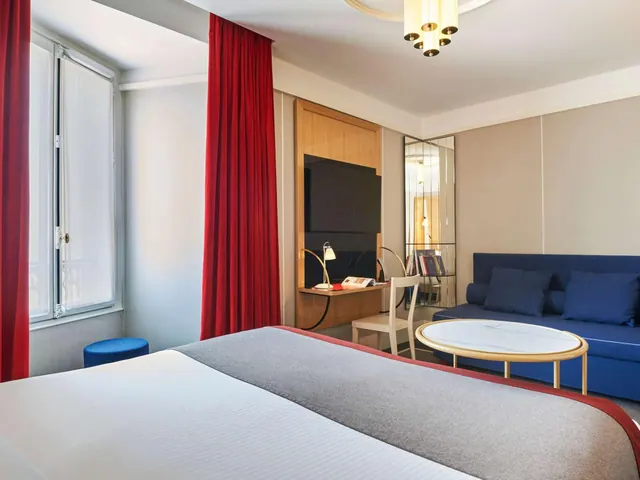 A bright hotel room contains a double bed, blue sofa, desk, television, and tall windows with red curtains.