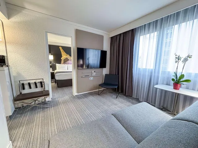 A bright hotel sitting area has a large sofa, television, armchairs and a window with sheer curtains beside a potted orchid.