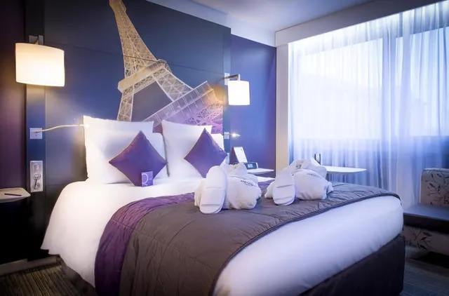 A modern hotel bedroom has a large bed, purple accents, Eiffel Tower wall artwork, and a seating area beside the window.