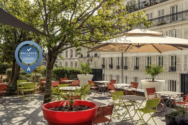 A furnished outdoor terrace with café tables, colorful chairs, umbrellas, potted plants, and a Parisian-style building beyond.