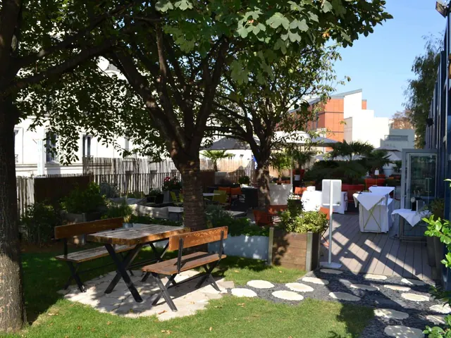 A landscaped outdoor dining terrace has wooden picnic tables, white-clothed tables, and mature trees.