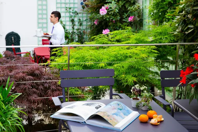 A furnished terrace surrounded by lush plants includes a table with an open book and fruit.