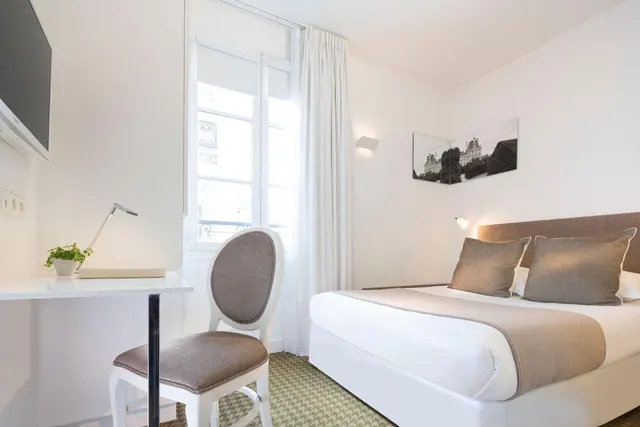 A bright hotel bedroom has a double bed, a compact desk, a television, and a tall curtained window.