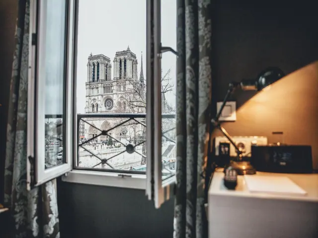 An open window looks toward Notre-Dame Cathedral from a room with a desk and lamp.