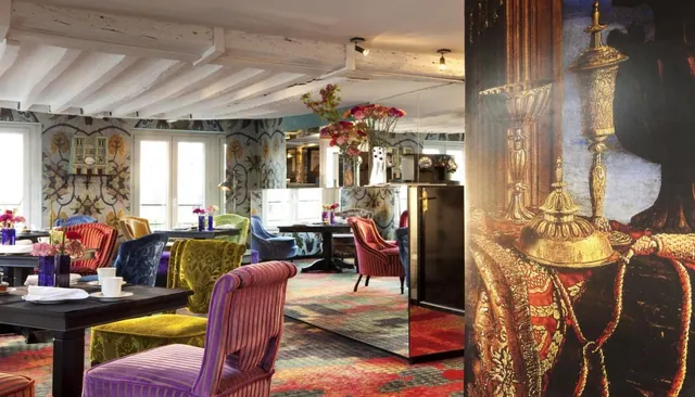 A colorful dining room contains patterned armchairs, set tables, exposed ceiling beams, and decorative wall artwork.