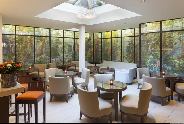 A bright hotel lounge contains upholstered chairs, sofas, small tables, floral window panels and a central skylight.