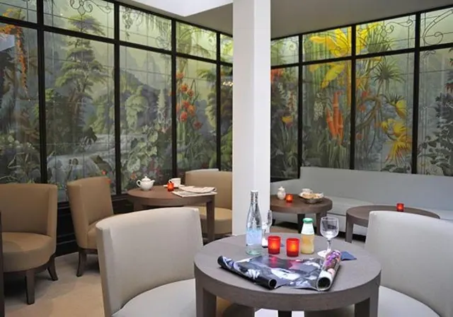A dining room with round tables, upholstered chairs, candles, and botanical mural panels on the windows.
