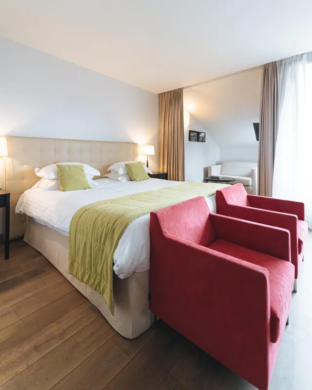 A bright hotel bedroom has a large bed, red armchairs, a sofa, and floor-to-ceiling curtains.
