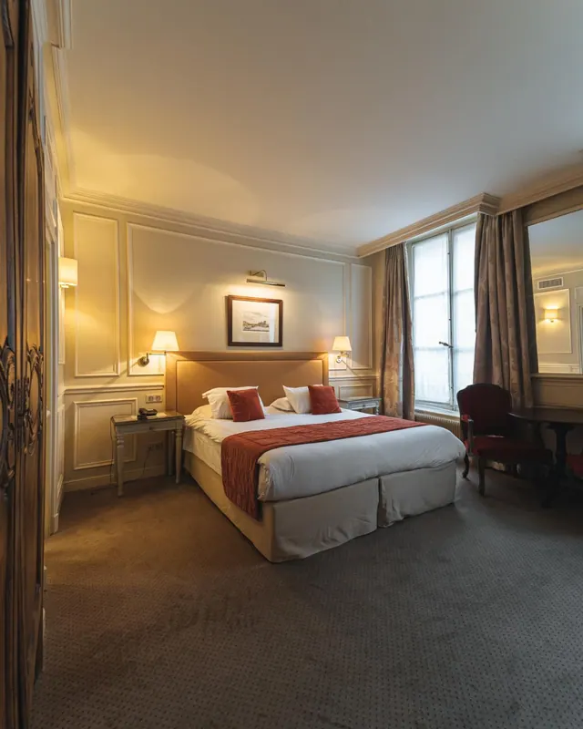 A classic hotel bedroom has a double bed, upholstered chair, desk and tall curtained windows.