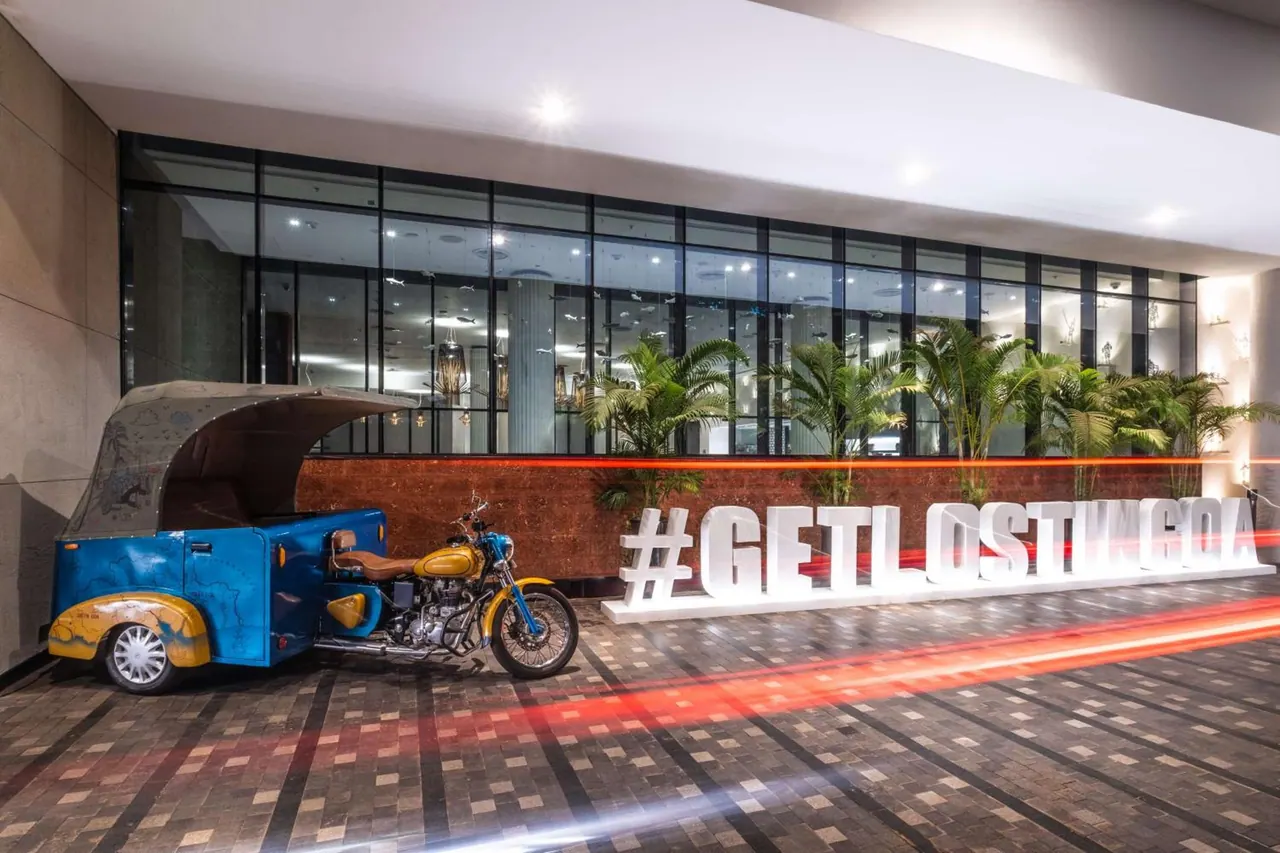 A brightly lit hotel entrance features a blue and yellow auto rickshaw beside large hashtag lettering and potted palms.