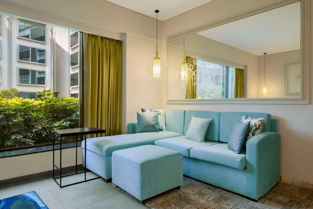 A bright living area contains a blue sectional sofa, ottoman, side table, large window and wall mirror.