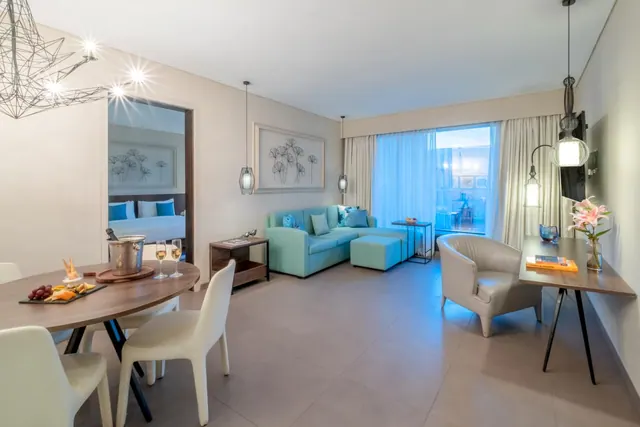 A bright modern living area has a sofa, dining table, desk, television and refreshments.