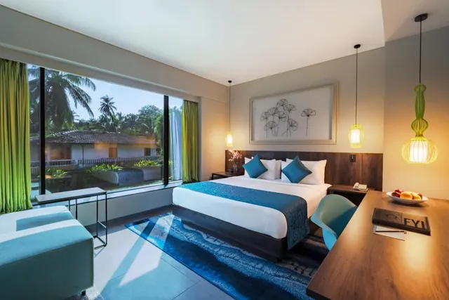 A bright modern bedroom has a double bed, desk, seating area and large windows overlooking palm trees and a garden.