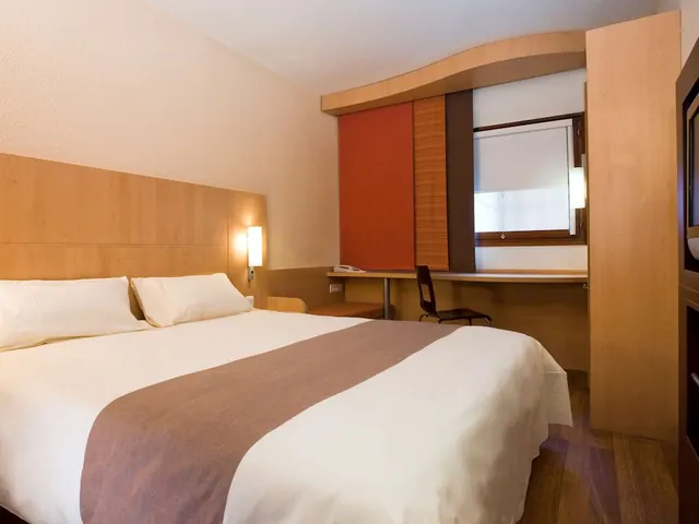 A double bedroom has a made bed, desk, wardrobe, television and window with a roller blind.