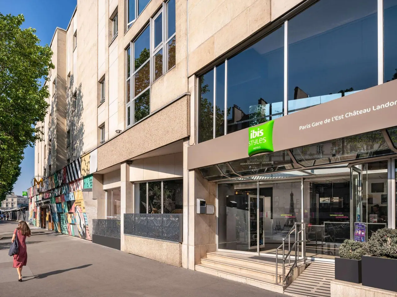 The hotel entrance has a glass canopy, steps, handrails and a green ibis Styles sign beside a city sidewalk.