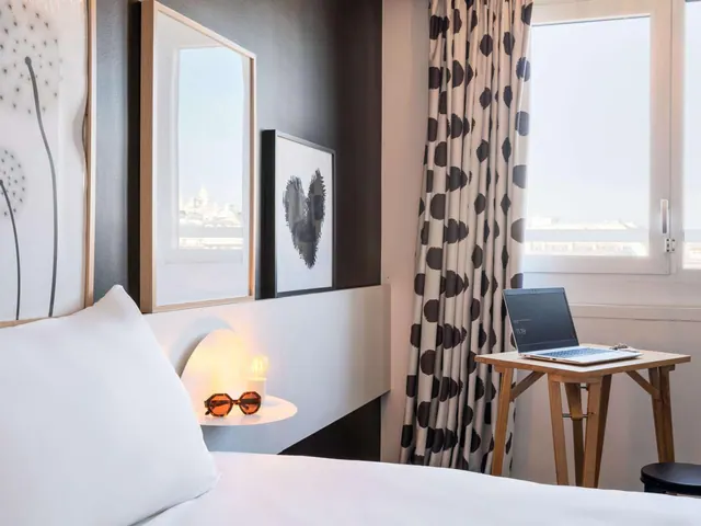 A bright hotel bedroom with a double bed, desk, patterned curtains and windows overlooking distant city buildings.