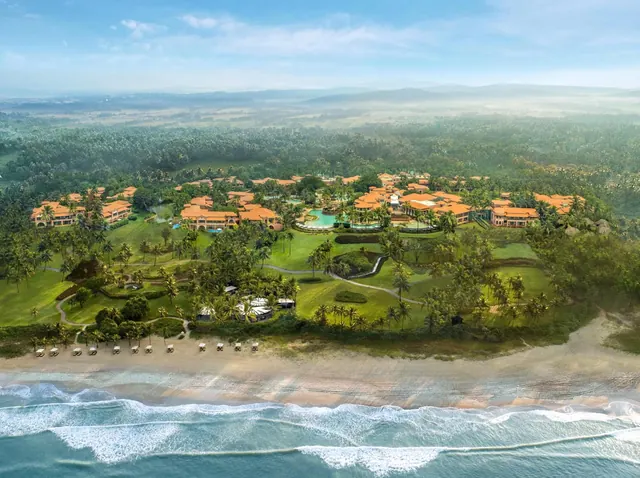 An aerial view shows a tropical resort with red-roofed buildings, landscaped grounds, a pool, and a sandy beach beside the sea.