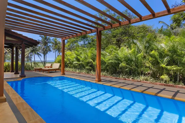 A large outdoor pool sits beneath a wooden pergola beside tropical gardens with the sea visible in the distance.