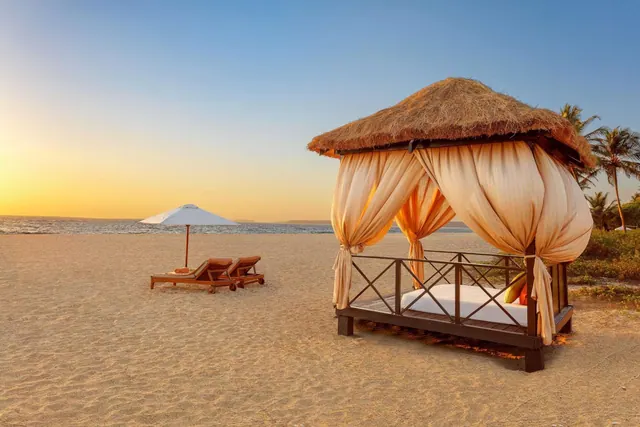 A sandy beach at sunset has a curtained thatched cabana, loungers, an umbrella and the sea beyond.